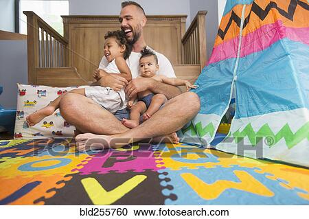 Stock Image - Father sitting on floor of playroom holding son and daughter. Fotosearch