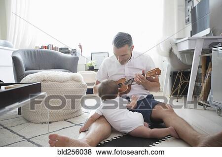 Stock Photo - Father sitting on floor playing ukulele for baby son. Fotosearch
