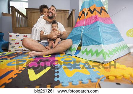 Stock Image - Father sitting on playroom floor holding son and daughter. Fotosearch