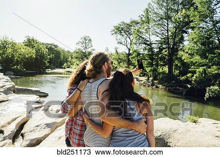 Stock Image - Friends posing for cell phone selfie on rocks near river. Fotosearch