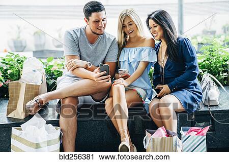 Stock Image - Friends sitting on bench texting on cell phones. Fotosearch