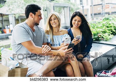 Stock Image - Friends sitting on bench texting on cell phones. Fotosearch