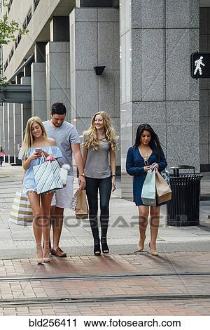 Stock Image - Friends walking in city carrying shopping bags texting on cell phones. Fotosearch