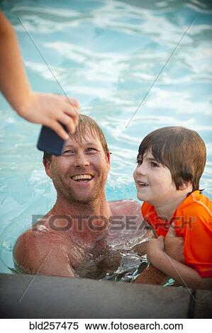 Stock Photography - Hand holding cell phone for Caucasian father and son in swimming pool. Fotosearch