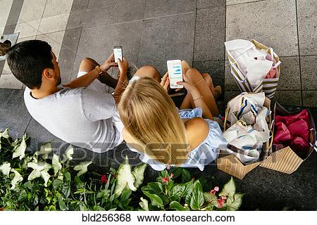 Stock Photo - High angle view of Caucasian couple with shopping bags texting on cell phones. Fotosearch