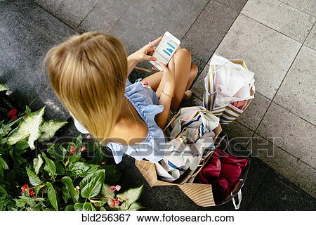 Stock Photo - High angle view of Caucasian woman with shopping bags texting on cell phone. Fotosearch