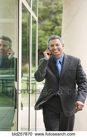 Stock Image - Hispanic businessman walking near window talking on cell phone. Fotosearch