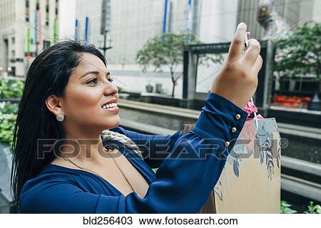 Stock Image - Hispanic woman with shopping bag posing for cell phone selfie. Fotosearch