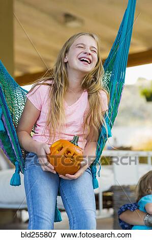 Stock Photo - Laughing Caucasian girl in hammock holding pumpkin. Fotosearch