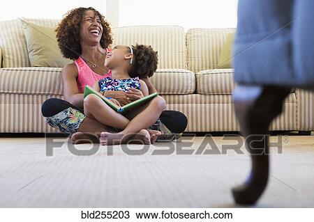 Stock Image - Laughing mixed race mother and daughter reading book on floor. Fotosearch