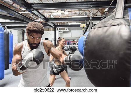 Stock Image - Man and women wearing boxing gloves hitting heavy bags. Fotosearch