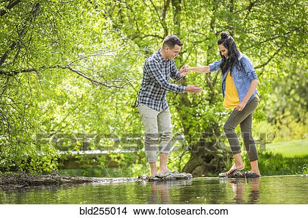 Picture - Man helping woman standing on rocks crossing river. Fotosearch