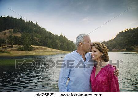 Stock Image - Man kissing woman on forehead near lake. Fotosearch
