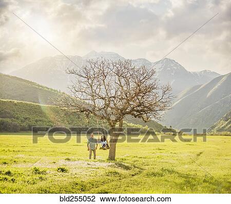 Stock Image - Man pushing woman swinging on tree swing. Fotosearch