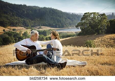 Picture - Man sitting on blanket in field playing guitar for woman. Fotosearch