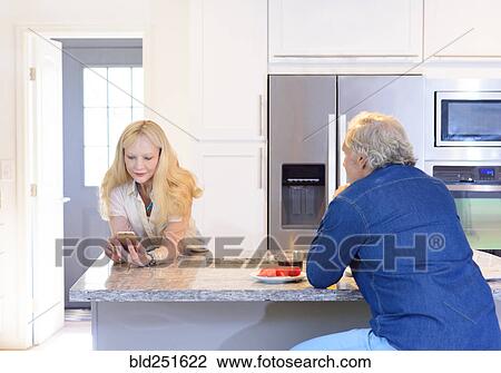 Stock Image - Man watching Caucasian woman texting on cell phone in kitchen. Fotosearch