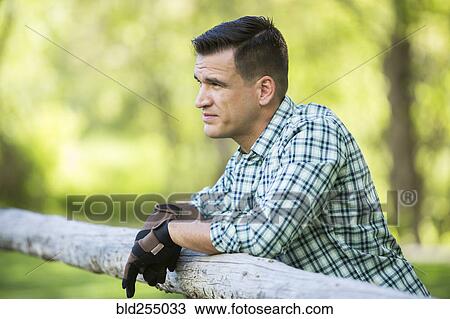 Stock Image - Man wearing gloves leaning on wooden fence. Fotosearch