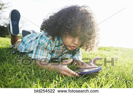 Stock Image - Mixed race boy laying in grass holding cell phone. Fotosearch