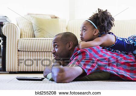 Stock Photo - Mixed race father and daughter laying on floor watching television. Fotosearch