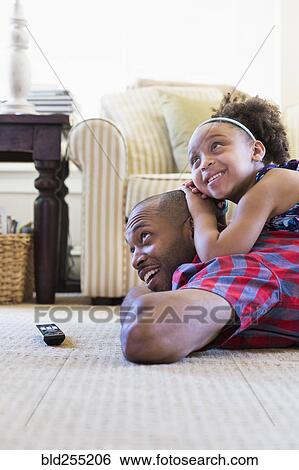 Stock Photograph - Mixed race father and daughter laying on floor watching television. Fotosearch