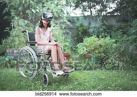 Picture - Mixed race girl in wheelchair using virtual reality goggles in backyard. Fotosearch