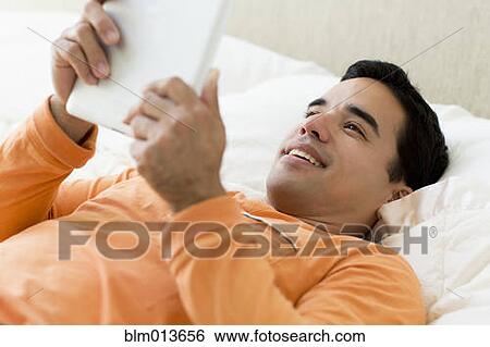 Stock Photograph - Mixed race man laying in bed using digital tablet. Fotosearch