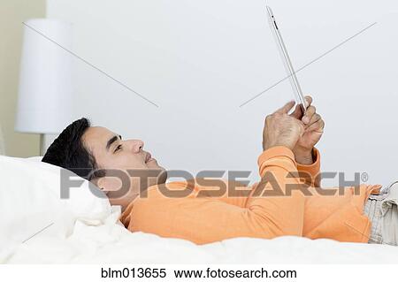 Stock Photography - Mixed race man laying in bed using digital tablet. Fotosearch