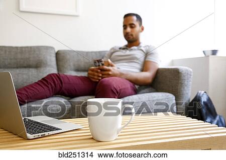 Picture - Mixed race man on sofa texting on cell phone near coffee and laptop. Fotosearch