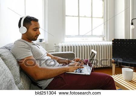 Picture - Mixed race man sitting on sofa listening to laptop with headphones. Fotosearch
