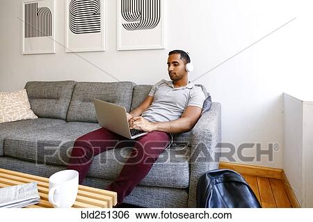 Mixed race man sitting on sofa listening to laptop with headphones View Large Photo Image Stock Photograph - Mixed race man sitting on sofa listening to laptop with headphones. Fotosearch
