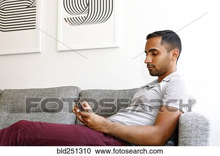 Stock Image - Mixed race man sitting on sofa texting on cell phone. Fotosearch