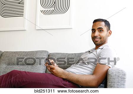 Stock Image - Mixed race man sitting on sofa texting on cell phone. Fotosearch