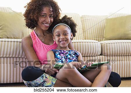 Picture - Mixed race mother and daughter reading book on floor near sofa. Fotosearch
