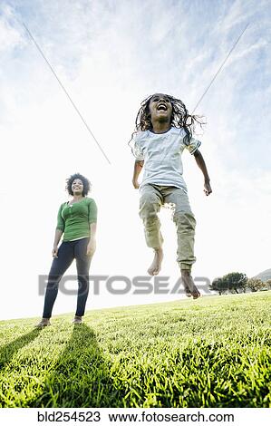 Stock Image - Mixed race mother watching daughter jumping in the grass. Fotosearch