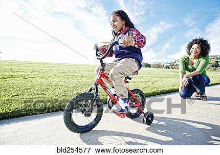 Stock Photo - Mixed race mother watching daughter riding bicycle. Fotosearch