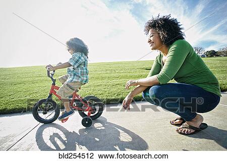 Stock Image - Mixed race mother watching son ride bicycle. Fotosearch