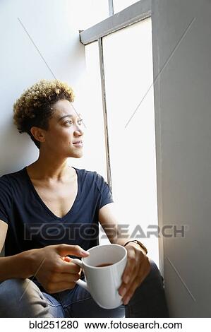 Stock Image - Mixed race woman drinking coffee on windowsill. Fotosearch