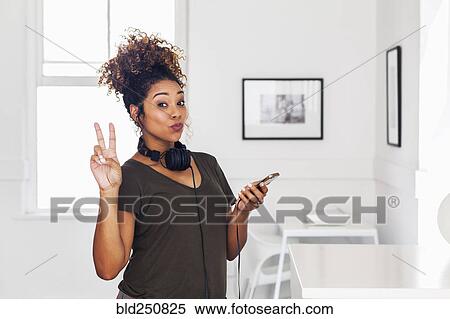 Stock Photography - Mixed race woman holding cell phone gesturing peace. Fotosearch