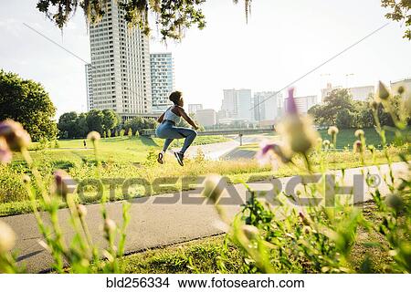 Picture - Mixed Race woman jumping on running path in park beyond wildflowers. Fotosearch