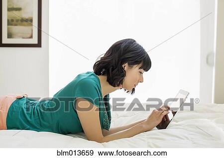 Stock Photo - Mixed race woman laying on bed using digital tablet. Fotosearch