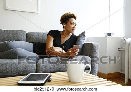 Stock Photo - Mixed race woman laying on sofa listening to earbuds. Fotosearch
