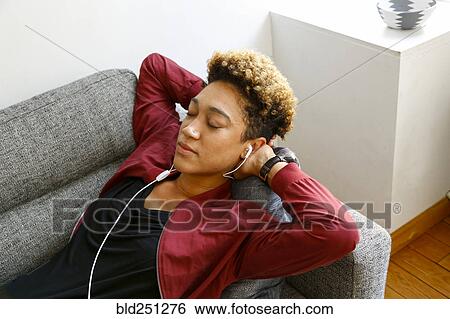 Stock Photograph - Mixed race woman laying on sofa listening to earbuds. Fotosearch