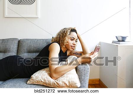 Stock Image - Mixed race woman laying on sofa texting on cell phone. Fotosearch