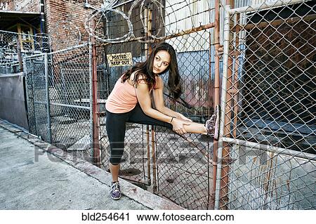 Stock Image - Mixed race woman leaning on chain link fence stretching leg. Fotosearch