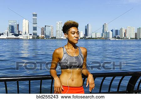 Stock Image - Mixed race woman leaning on railing at waterfront listening to earbuds. Fotosearch