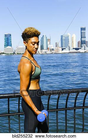 Stock Image - Mixed race woman lifting dumbbells at waterfront. Fotosearch