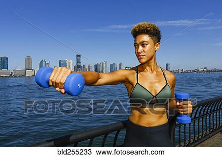 Stock Image - Mixed race woman lifting dumbbells at waterfront. Fotosearch