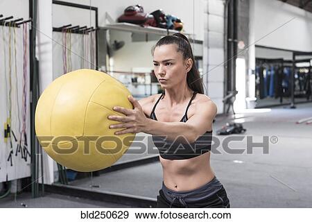 Stock Photo - Mixed race woman lifting heavy ball in gymnasium. Fotosearch