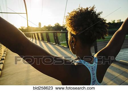 Stock Image - Mixed Race woman wearing earbuds stretching arms on bridge. Fotosearch