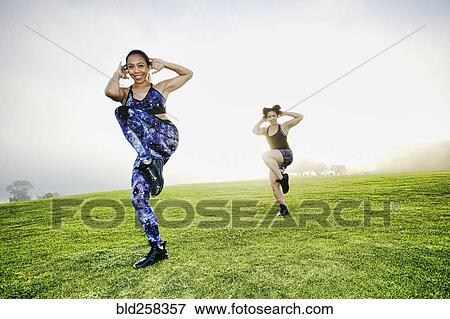 Stock Photo - Mixed race women standing on one leg in field. Fotosearch